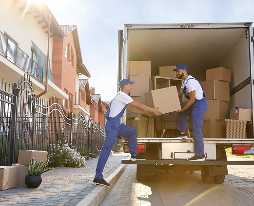Cartons Déménagement Salon Ancien Lumière Naturelle Gros cartons de déménagement scellés au premier plan dans une pièce peu éclairée avec fenêtre et un canapé bleu et jaune visible au fond.