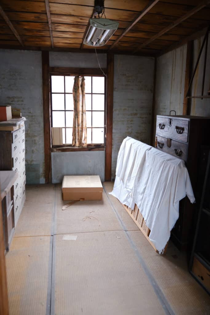 Pièce japonaise ancienne : meubles, tatamis et lumière tamisée Intérieur sombre d'une pièce japonaise ancienne avec plafond en bois, murs patinés, tatamis au sol et une fenêtre grillagée. Des meubles anciens et un carton « 座布団 » sont visibles.