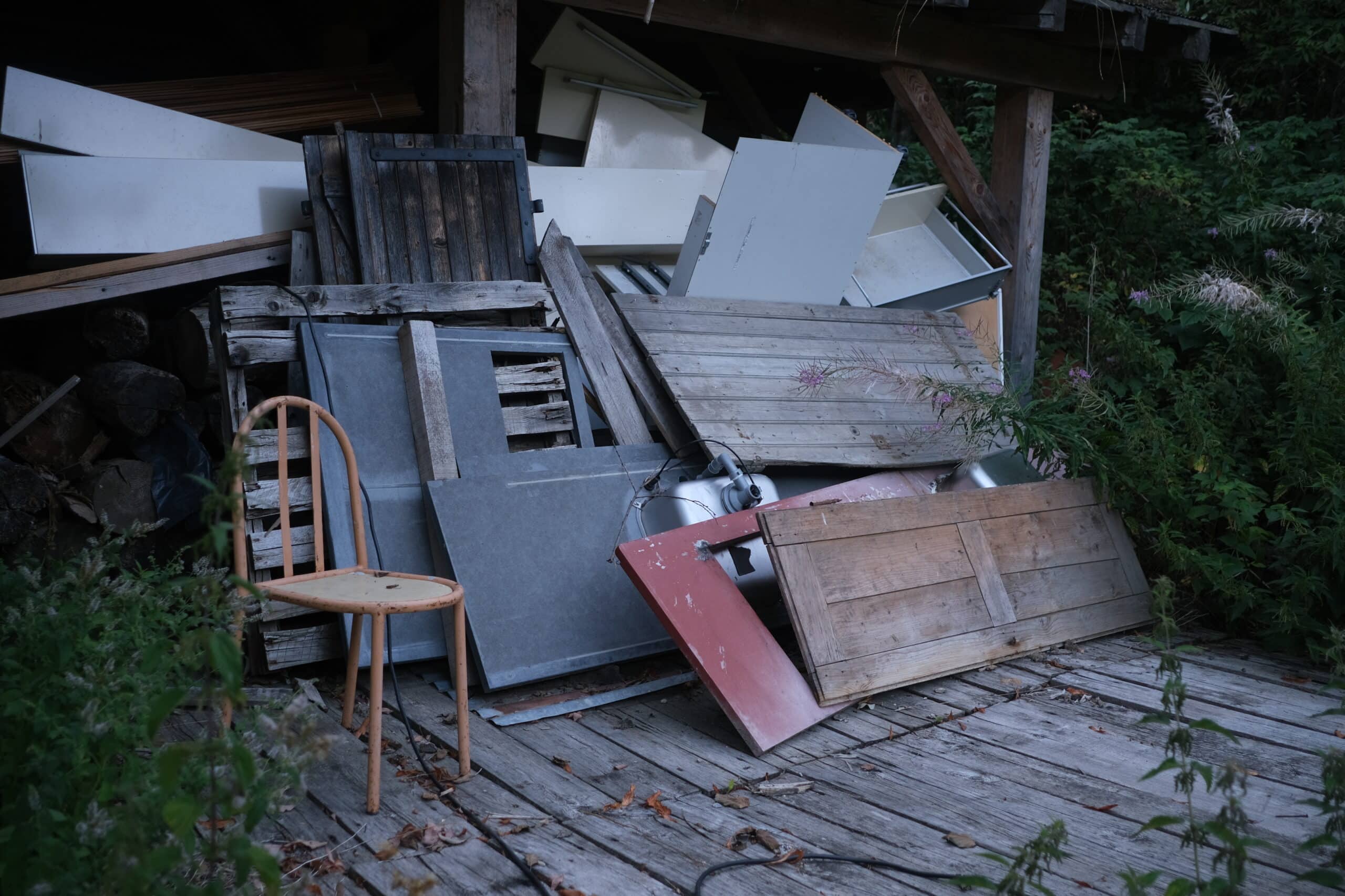 Vieux objets et débris, incluant des panneaux, bois empilés, et une chaise orange délavée sur un plancher de bois grisonnant près d'une végétation dense.